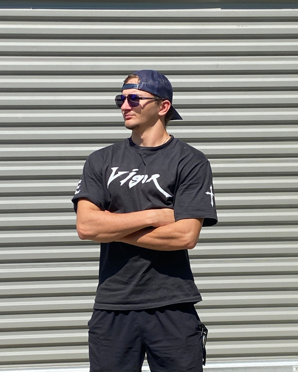 Person wearing a black t-shirt with white text and black pants against a corrugated metal wall.