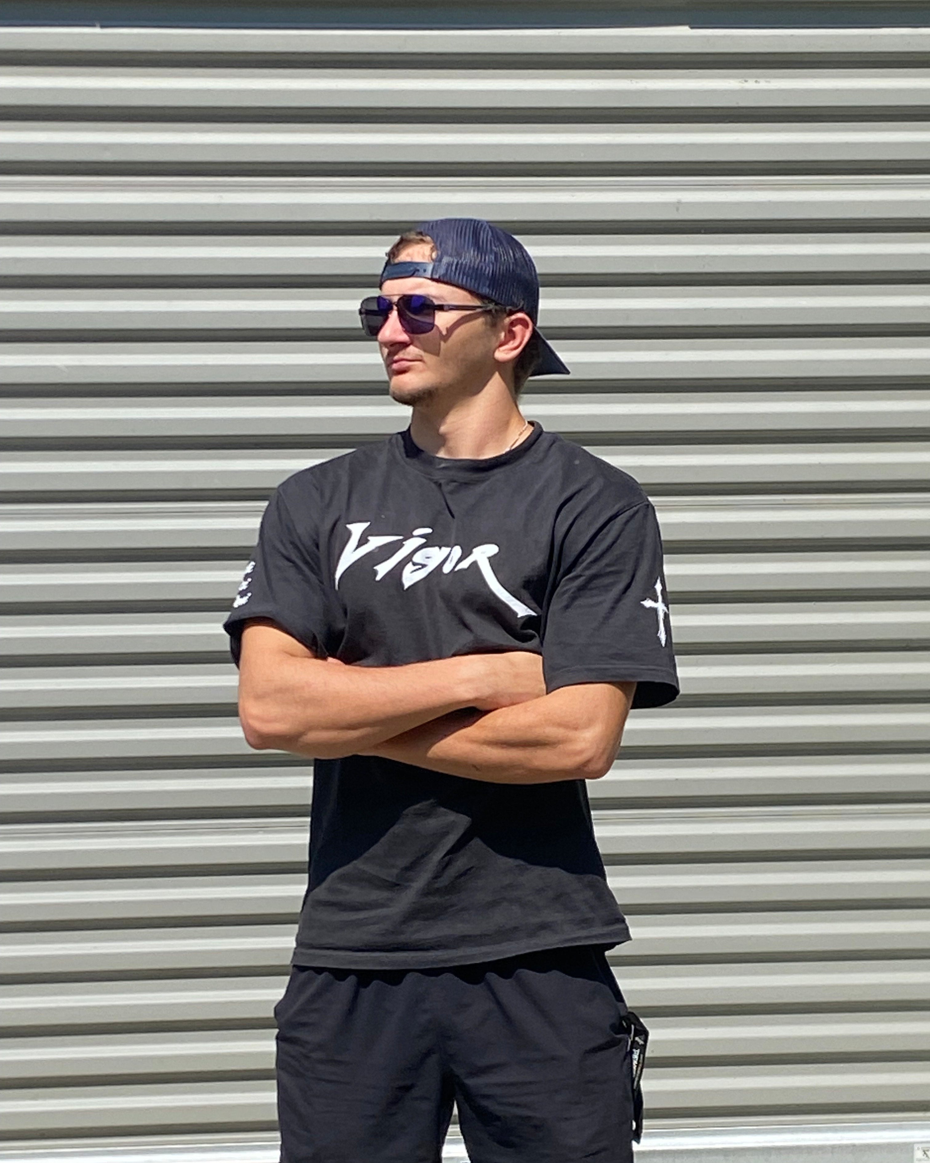 Person wearing a black t-shirt with white text and black pants against a corrugated metal wall.